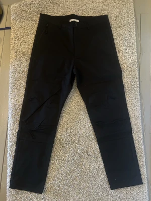 Svarta  byxor från Stone Island - Svarta byxor från Stone Island med klassisk logotyp på bakfickan. Toppen passform.. Byxorna har dragkedja och knapp framtill samt fickor med dragkedja på sidorna. Perfekt för dig som gillar stilrena och snygga streetwear-plagg.