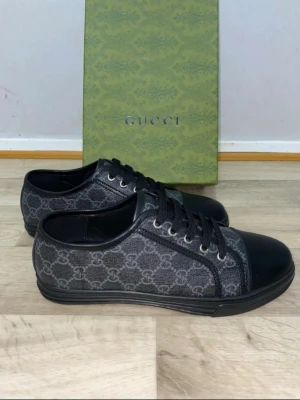 Svarta sneakers från Gucci med GG-mönster - Snygga svarta sneakers från Gucci med klassiskt GG-mönster i grått. Skorna har rund tå, platt sula och snörning framtill. Materialet är en mix av skinn och textil, vilket ger en lyxig känsla. Perfekta för dig som gillar exklusiva märken och stilren design.