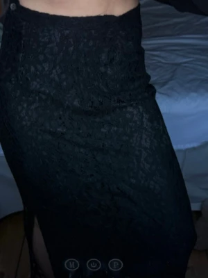 Svart spetskjol med slits - Säljer en svart långkjol i spets med snygg slits på sidan och dragkedja i midjan. Kjolen har en tight passform och det genombrutna spetsmaterialet ger en cool och elegant vibe. Perfekt att styla med boots eller klackar för en trendig look.