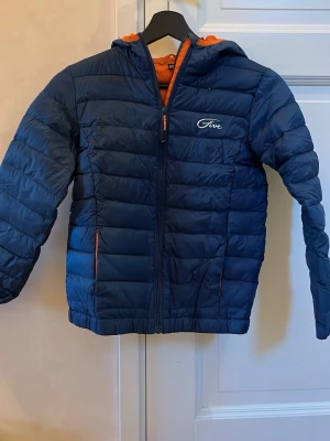 Mörkblå pufferjacka från Five - Säljer en mörkblå pufferjacka från Five med orange foder och huva. Jackan har dragkedja framtill och två fickor med orange detaljer. Perfekt för kyliga dagar och har en snygg, quiltad look.