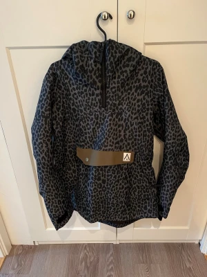 Leopardmönstrad vindjacka med huva - Snygg vindjacka från Arctic Legacy i mörkgrått med svart leopardmönster. Jackan har huva, half zip framtill och praktiska fickor med dragkedja. Materialet är polyester och jackan har en loose passform. Perfekt för dig som gillar att sticka ut med coola mönster.