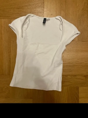 Vit ribbad topp från H&M - Säljer en enkel och stilren vit topp från H&M med ribbad struktur. Toppen har korta ärmar och en fyrkantig ringning som ger en modern look. Perfekt att matcha med jeans eller kjol för en clean outfit.