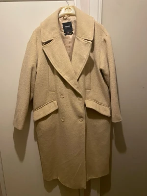 Beige kappa från Pull&Bear - Snygg beige kappa från Pull&Bear med bred krage, dubbelknäppning och två stora fickor framtill. Jackan har lång ärm och en stilren slits bak. Materialet är mjukt och känns lyxigt, perfekt för dig som vill ha en clean look.