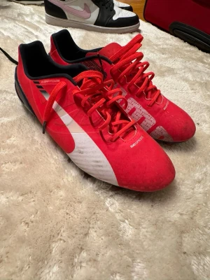 Röda Puma EvoSpeed fotbollsskor - Säljer ett par röda Puma EvoSpeed fotbollsskor med svart och vit detalj, snörning och mönstrad ovansida med stora Puma-loggor. Skorna har dobbar för gräsplan och är tillverkade i syntetmaterial. 