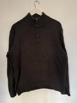 Grå half zip tröja från Polo Ralph Lauren - Mörkgrå stickad tröja från Polo Ralph Lauren med ribbad hög krage och half zip-dragkedja. Broderad röd logga på bröstet. Tillverkad i 100% bomull och har långa ärmar samt ribbade muddar för en clean look.