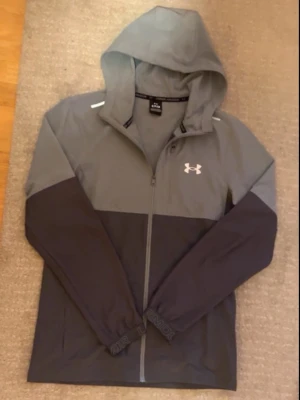 Grå vindjacka från Under Armour - Snygg tvåfärgad vindjacka från Under Armour i ljusgrått och mörkgrått. Jackan har huva, hel dragkedja framtill och elastiska muddar med logga. Perfekt för träning eller när du vill ha en sportig look. Tillverkad i lätt polyester som andas.