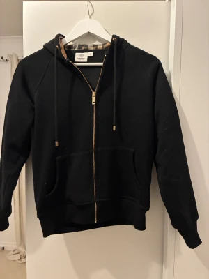 Svart Burberry hoodie med dragkedja - Svart hoodie från Burberry med klassiskt rutigt foder i huvan, guldfärgad dragkedja och snören med metalländar. Mjuk bomullskänsla, två fickor fram och snyggt Burberry-märke i nacken. Perfekt för dig som gillar stilrena och lyxiga detaljer.