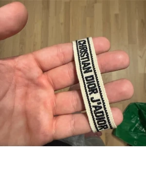 Beige Christian Dior J'Adior västband - Snyggt västband från Christian Dior med texten 'J'Adior' i svart på beige bakgrund. Bandet är vävt och har dekorativa tofsar i ändarna, perfekt för att ge din väst en lyxig touch. Stilren design med svarta linjer längs kanterna.