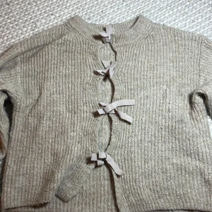 Beige stickad kofta från Gina Tricot - Säljer en beige stickad kofta från Gina Tricot i storlek 146/152. Koftan har långärmade ärmar och snygga knytband framtill som ger en cool touch. Perfekt för dig som gillar mjuka och mysiga plagg med lite extra detaljer.