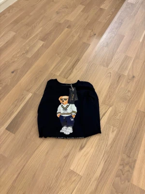 Mörkblå sweatshirt Polo Bear Ralph Lauren - Snygg mörkblå sweatshirt från Ralph Lauren med Polo Bear-tryck framtill. Tröjan har rund hals, ribbade muddar och är långärmad. Materialet är mjuk bomull och polyester, perfekt för en chill och stilren look. Taggarna sitter kvar.