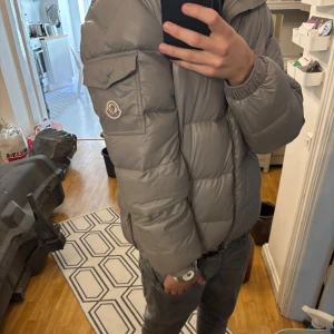 Grå Moncler pufferjacka med luva - Säljer en snygg grå pufferjacka från Moncler med luva och dragkedja framtill. Jackan har Moncler-logga på ärmen, praktiska fickor och quiltad design. Nfc till jackan funkar. Perfekt för kalla dagar och riktigt clean look. Hör gärna av er vid mer frågor, pris kan diskuteras!😁