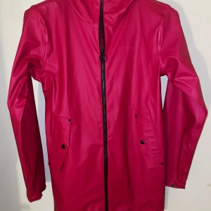 Rosa regnjacka från Didriksons - Snygg rosa regnjacka från Didriksons i Galon-material, med huva och hel dragkedja framtill. Jackan har två stora fickor med knappar och justerbara ärmslut. Perfekt när du vill hålla dig torr och ändå se cool ut.