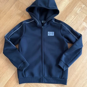 Svart hoodie från EA7 Emporio Armani - Svart hoodie från EA7 Emporio Armani med dragkedja framtill och huva. Snygga vita detaljer längs ärmarna och logga på bröstet. Har lite små slitningar här och där som knappt syns för andra när du har på dig den 
