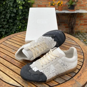 Maison Margiela sneakers  - Maison Margiela sneakers i vitt skinn och mocka med unika svarta färgstänk och beige snörning. Snygg kontrast mellan den ljusa basen och den mörkblåa tån. Klassisk siluett med gummisula och diskreta detaljer. Kommer med originalkartong.