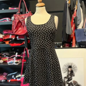 Svart prickig kortklänning H&M - Svart kortklänning från H&M med vita prickar, rundad hals och djup rygg. Klänningen är ärmlös och har en lätt utställd kjol. Materialet är mjuk och stretchig polyester med elastan, vilket gör den bekväm och följsam.