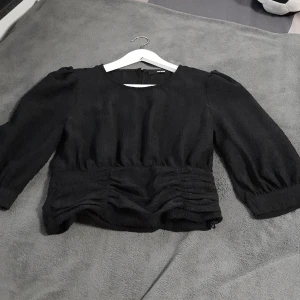 Svart blus med puffärm från BikBok - Säljer en svart blus från BikBok i storlek S. Blusen har puffiga trekvartsärmar, rund halsringning och snygga rynkade detaljer vid midjan. Materialet är strukturerat och ger en cool känsla. Perfekt att styla med jeans eller kjol.