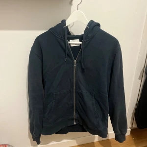 Mörkblå hoodie med dragkedja från Colorful Standard - Mörkblå hoodie från Colorful standard med dragkedja framtill, huva med snörning och två stora fickor. Tröjan är i mjuk bomull och har en avslappnad look. Den har täcken på användning och ett av snörena är lite sönder. Övrigt är skicket 6/10
