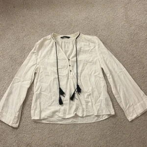 Vit tröja från Zara med snörning - Säljer en vit tröja från Zara i storlek S. Tröjan har långa ärmar, knappar framtill och cool snörning med svarta tofsar vid halsen. 