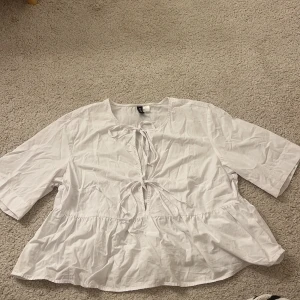 Vit blus med snörning från H&M - Säljer en vit blus från H&M Divided i storlek M. Blusen har korta ärmar, volang nedtill och två snörningar framtill som ger en söt och trendig look. Aldrig använd med prislappen är inte kvar!