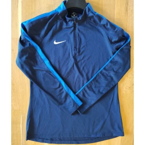 Nike Dri-Fit blå långärmad träningströja - Nike Dri-Fit långärmad träningströja i marinblått med ljusblå detaljer längs ärmarna och vit broderad swoosh på bröstet. Tröjan har half zip framtill och är tillverkad i ett lätt, syntetiskt material som andas. Perfekt för träning eller sport.