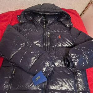 Marinblå pufferjacka från Polo Ralph Lauren - Säljer en marinblå glansig pufferjacka från Polo Ralph Lauren med röd logga på bröstet. Jackan har huva, flera dragkedjeförsedda fickor och quiltad design. Perfekt för kalla dagar och riktigt snygg till streetwear-stilen.
