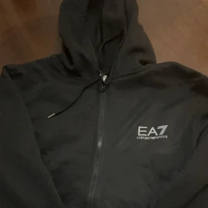 Svart EA7 hoodie med dragkedja - Svart hoodie från EA7 Emporio Armani med vit logga på bröstet. Tröjan har huva med snörning, hel dragkedja framtill och långa ärmar. Perfekt för en chill och sportig look. Helt ny och använd 