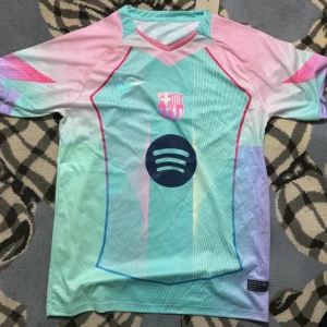 Barcelona fotbollströja Nike pastell - Säljer en Barcelona fotbollströja från Nike i pastellfärger med klubbmärke och Spotify-logga på bröstet. Tröjan har korta ärmar och V-ringad hals. Färgerna är mintgrön, rosa och ljuslila med grafiska detaljer och tunna linjer. Perfekt för dig som gillar unik design.