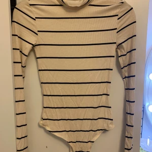 Randig polotopp i beige och svart - Säljer en beige polotopp med svarta horisontella ränder. Toppen är ribbad och har lång ärm samt hög krage. Materialet känns mjukt och stretchigt, perfekt för att styla med jeans eller kjol. Kroppsnära passform och klassisk look.