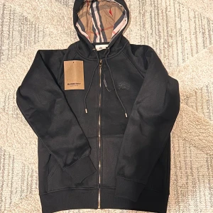 Svart hoodie från Burberry med dragkedja - Helt ny kommer med påse och ett plagg som du ser på första bilden .Svart hoodie från Burberry med klassiskt rutigt foder i huvan, guldfärgad dragkedja och snörning. Tröjan har långa ärmar, ribbade muddar och två fickor framtill. Perfekt för dig som vill ha en stilren och lyxig look.