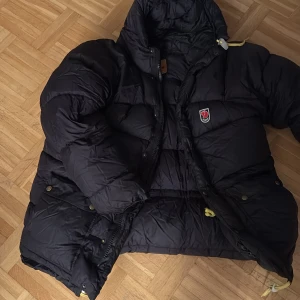 Svart dunjacka från Fjällräven - Säljer en svart dunjacka från Fjällräven med huva och gula detaljer. Jackan har klassisk puffer-design, dragkedja och tryckknappar framtill samt två stora fickor. Perfekt för kalla dagar och riktigt varm.