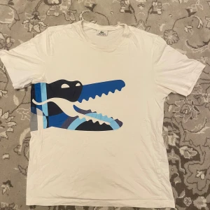 Vit Lacoste t-shirt  - Säljer en stilren vit t-shirt från Lacoste Sport med en stor, grafisk krokodil i blå och svart på framsidan. Inga direkta defekter, behöver bara styrkas. Storlek 5 vilket jag skulle säga motsvarar en M.