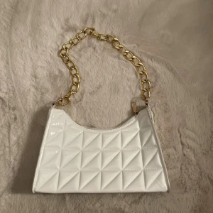Axelväska med guldfärgad kedja - Säljer en superfin vit axelväska med guldfärgad kedja – perfekt accessoar för både vardag och fest! Den är använd 1 gång.