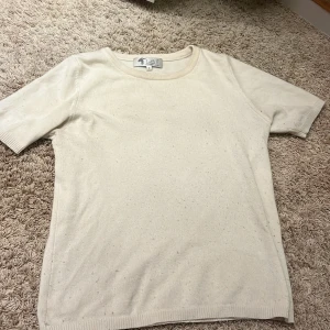 Beige kashmir kortärmad tröja från Lotti P - En enkel och stilren beige kashmir tröja från Lotti P. Tröjan har korta ärmar, rund halsringning och är gjord i ett mjukt material som känns skönt mot huden. Perfekt att matcha med jeans eller kjol för en clean look.