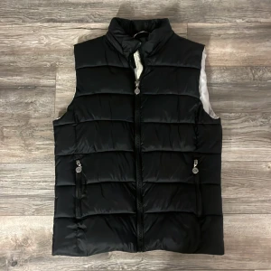 Svart dunväst från Colmar Originals - Svart dunväst från Colmar Originals med quiltad design och hög krage. Västen har två fickor med dragkedja framtill och en diskret logga på ryggen. Materialet är polyester och dun, perfekt för kyliga dagar. Stängs med dragkedja och har en stilren look. Den har en jette litet hål på in sidan men synts från yttre. Den passar en medium skulle jag säga en liten large