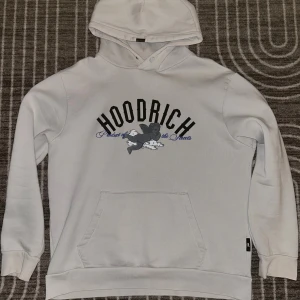Vit hoodie från Hoodrich med tryck - Säljer en vit hoodie från Hoodrich med svart/grå och blått tryck framtill och stort motiv med änglar och bokstaven H på ryggen. Tröjan har huva, känguruficka samt gömd ficka vid armen. Perfekt för dig som gillar streetwear och snygga detaljer. Obs säljs inte längre utav vad ja sätt via nätet!!