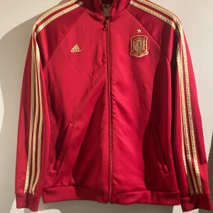 Röd Adidas Spanien track jacket - Röd track jacket från Adidas med Spaniens landslagsemblem och guldiga detaljer. Klassiska tre ränder längs ärmarna, dragkedja framtill och ståkrage. Perfekt för dig som vill ha sportig och snygg stil med fotbollsvibe. Pris kan diskuteras!! ❤️❤️❤️hör av er vid frågor! 