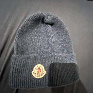 Svart ribbstickad mössa från Moncler - Ribbstickad svart mössa från Moncler med klassisk uppvikt kant och ikonisk Moncler-logga framtill. Mjuk och varm, perfekt för kyliga dagar. Enkel och stilren design som funkar till de flesta outfits.