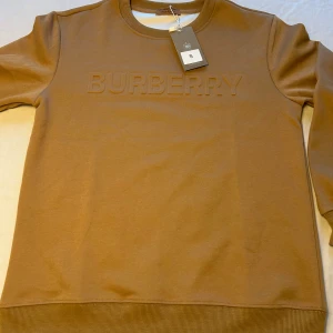 Beige sweatshirt  Burberry S - Snygg beige sweatshirt  Burberry med broderad logga framtill. Tröjan har rund halsringning, ribbade muddar och är tillverkad i en mjuk bomulls- och polyesterblandning. Perfekt för en clean och stilren look.