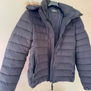 Svart pufferjacka från Superdry - Svart pufferjacka från Superdry med avtagbar huva och fuskpälsdetalj. Jackan har flera dragkedjefickor, hög krage och horisontella sömmar. Klassisk Superdry-logga på ärmen. Längd från axel oc ner ca 58 cm. Liten i storleken, mer som 36