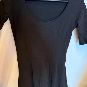 Svart kortärmad klänning i bomull - En enkel svart klänning med rundad hals och korta ärmar. Modellen är figurnära upptill och har en lätt utställd nederdel. Tillverkad i mjuk bomull som känns skön mot huden. Perfekt för dig som gillar minimalistisk stil och vill ha en klassisk look.