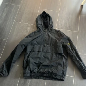 Svart vindjacka från Lyle & Scott  - Svart vindjacka från Lyle & Scott med huva och halv dragkedja. Jackan har elastiska muddar, stor ficka framtill och den klassiska gula loggan på ärmen. Tillverkad i lätt polyester, perfekt för blåsiga dagar. Ficka på ärmen också. Köpt för 799 