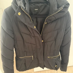 Svart pufferjacka från Oasis med huva - Säljer en svart pufferjacka från Oasis med guldig dragkedja och knappar. Jackan har huva, två fickor med dragkedja och snygg quiltad design. Perfekt för kyliga dagar och riktigt skön att bära. Passar dig som gillar stilren och enkel look.