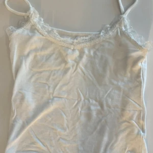 Vit linnetopp med spetskant - Säljer en söt vit linnetopp med tunna axelband och dekorativ spetskant längs halsringningen. Toppen är ärmlös och har en mjuk, lätt känsla som passar perfekt till varma dagar. Enkel och stilren design med romantisk touch.