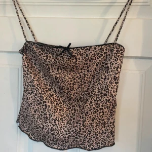 Leopardmönstrad linnetopp från Bershka - Säljer en snygg leopardmönstrad linnetopp från Bershka i storlek XS. Toppen har tunna axelband och en liten svart rosett framtill. Färgen är beige med svarta leopardfläckar och materialet känns lent och lätt mot huden. Perfekt för dig som gillar djurmönster!
