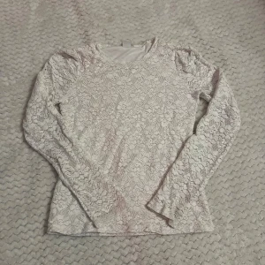 Vit långärmad spetsblus - Säljer en vit långärmad blus med blommigt spetsmönster. Toppen har rund hals och är figurnära i modellen, perfekt för dig som gillar romantisk stil. Materialet är mjukt och stretchigt, vilket gör den bekväm att bära hela dagen.