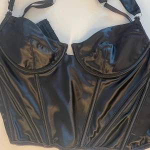 Svart korsett från her[body] XS - Snygg svart korsett från her[body] i storlek XS. Glansigt material med markerade sömmar och kupor som ger en cool siluett. Justerbara axelband för perfekt passform. Perfekt för dig som vill ha en statement topp med edge.