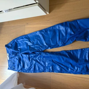 Blåa skinnbyxor från Bershka - Snygga blåa skinnbyxor från Bershka i storlek S. Byxorna har klassisk femficksmodell, bälteshällor och dragkedja fram. Materialet är glansigt och ger en cool look. Sitter riktig fint i passform som ett par jeans med stretch 