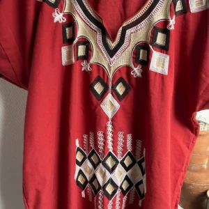 Röd klänning med broderade detaljer - Unik röd klänning med snygga broderade mönster i svart, beige och vitt. Klänningen har lös passform och geometriska broderier framtill som ger en cool och bohemisk vibe. Perfekt för dig som gillar statement-plagg med etnisk touch.