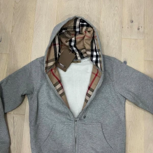 Grå Burberry hoodie med rutigt foder - Säljer en grå hoodie från Burberry med dragkedja och klassiskt rutigt foder i huvan. Tröjan har snörning i huvan, broderad logga på bröstet och två fickor framtill. Perfekt för dig som vill ha en stilren och lyxig streetwear-look.