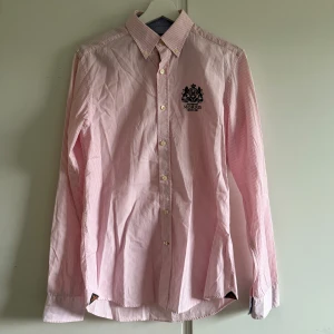 Rosa och vit randig skjorta Morris S - Snygg rosa och vit randig skjorta från Morris med broderad logga på bröstet. Klassisk button-down krage, långa ärmar och vita knappar framtill. Skjortan är gjord i mjuk bomull och har en stilren, normal passform. Perfekt för dig som gillar preppy stil och detaljer.
